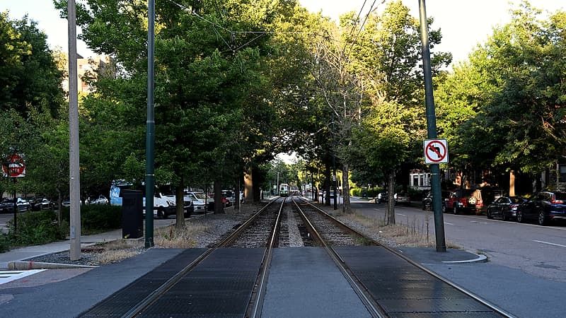 A rural road approaching a single track crossing with tall grass and brush growing close to the rail line