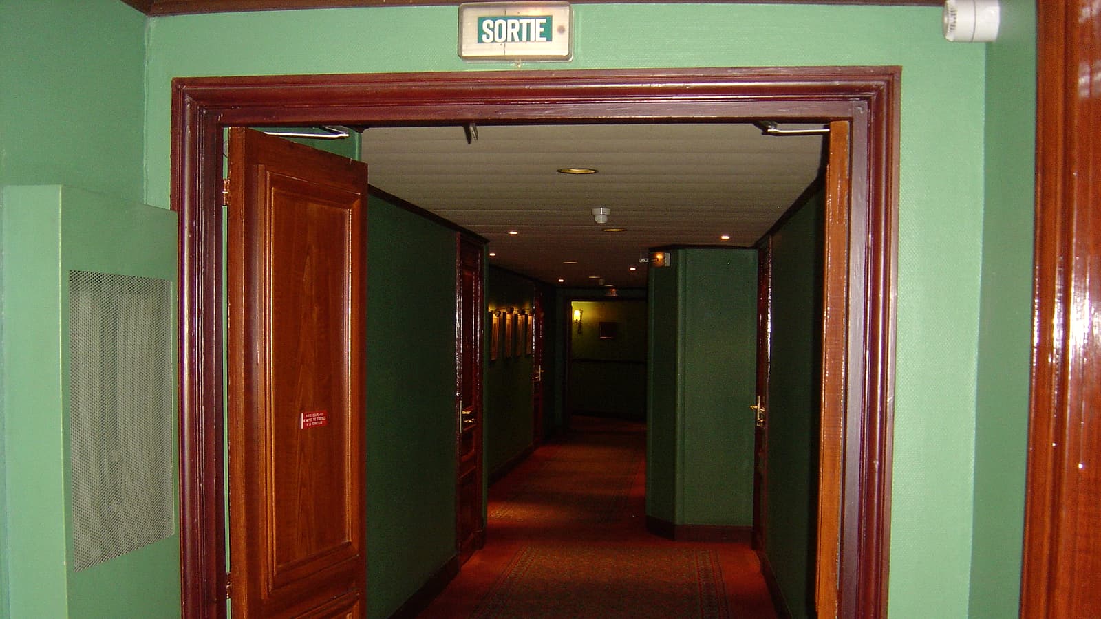 An empty hotel corridor with patterned carpet, closed numbered doors and wall sconces receding toward a window at the end