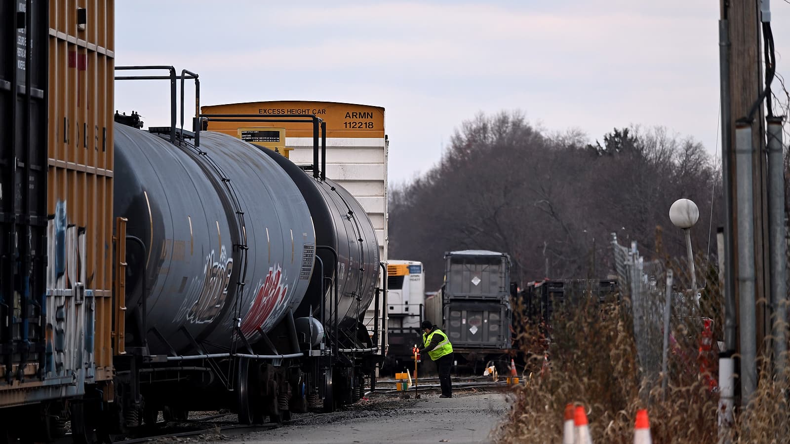 A railroad worker in a high visibility vest and hard hat standing beside freight cars in a rail yard at dusk